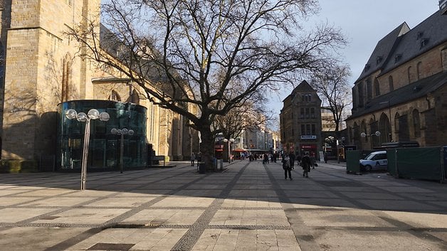 Eine breite Straße mit Fußgängern, flankiert von Gebäuden und einem großen Baum, unter klarem Himmel.