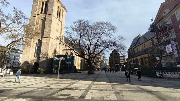 Ein sonniger Stadtplatz mit einer historischen Kirche, Bäumen und Passanten.