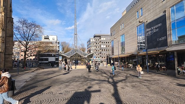 Une place urbaine animée avec des gens marchant, des magasins et une structure en verre sous un ciel bleu.