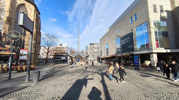 Ein belebter Fußgängerplatz mit Geschäften und Cafés, links eine Kirche und viele Menschen, die gehen.