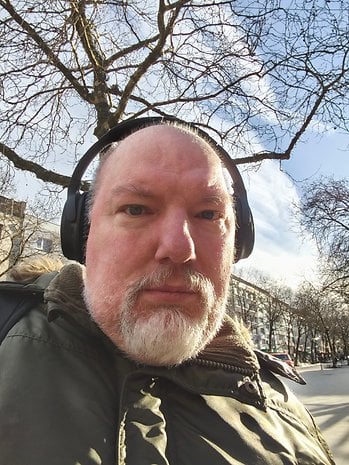 Un homme avec des écouteurs, portant une veste verte, se tient dehors sous un arbre avec un ciel dégagé en arrière-plan.