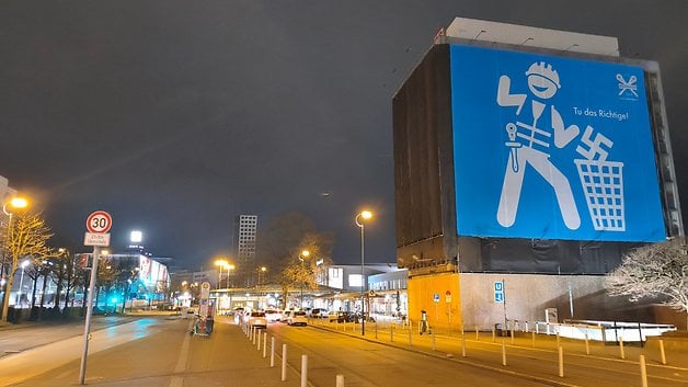 Un grand panneau bleu affichant un travailleur cartoon et le texte 'Tu das Richthigel' de nuit.