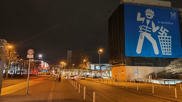 Une rue la nuit avec des panneaux de circulation et une grande bannière bleue représentant un personnage de dessin animé tenant des outils.