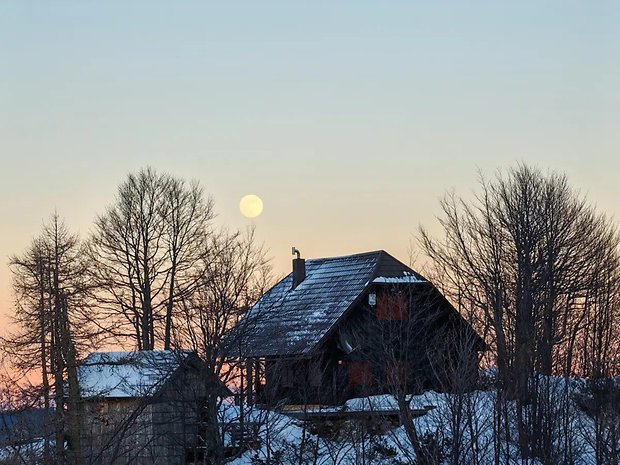 Eine verschneite Landschaft mit einer dunklen Holzhütte und einem vollen Mond, der hinter Bäumen bei Sonnenuntergang aufgeht.