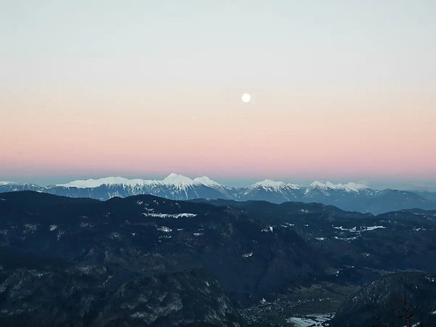 Eine ruhige Berglandschaft bei Dämmerung mit einem vollen Mond und pastellfarbenem Himmel.