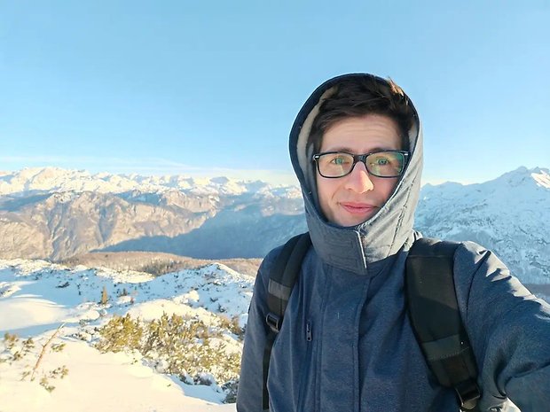 Eine Person in einer blauen Jacke mit Brille steht in einer schneebedeckten Berglandschaft unter einem klaren Himmel.