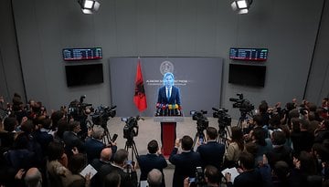 A press conference in Albania with a speaker at a podium, flanked by cameras and an Albanian flag.