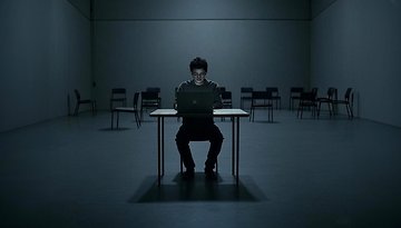 Eine Person sitzt allein an einem Tisch mit einem Laptop in einem dunklen, leeren Raum.