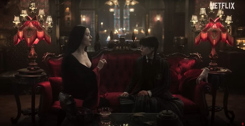 Morticia Addams sitzt auf einer roten Couch und spricht mit Wednesday in einer gotischen Umgebung. Netflix-Logo sichtbar. Morticia Addams sitzt auf einer roten Couch und spricht mit Wednesday in einer gotischen Umgebung. Netflix-Logo sichtbar.
