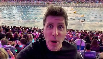 A man takes a selfie in a crowded arena during a swimming event, with colorful lights in the audience.