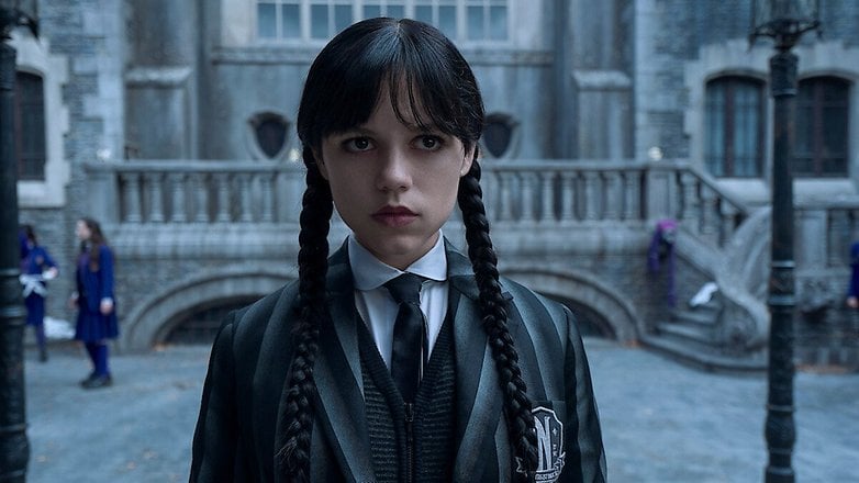 Wednesday Addams steht in einem dunklen Outfit vor einem gotischen Herrenhaus und sieht ernst aus. Wednesday Addams steht in einem dunklen Outfit vor einem gotischen Herrenhaus und sieht ernst aus.