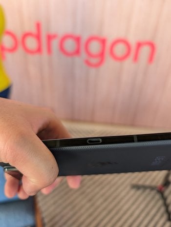 En person som håller en Asus ROG Phone 9, som visar USB-C-porten, med en suddig Snapdragon-logotyp i bakgrunden.