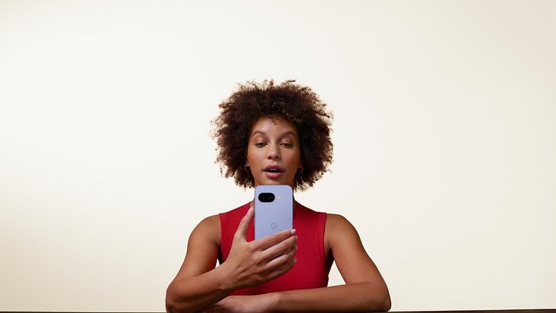 A person holds a Google Pixel 9a phone, looking at it while seated at a table against a light background.