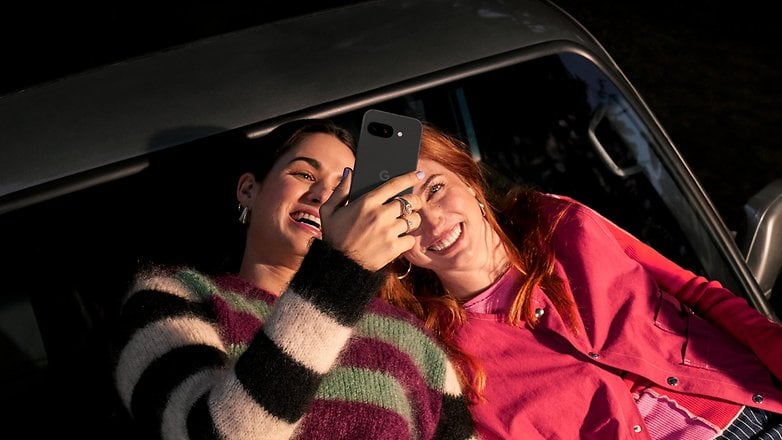 Two women laughing and looking at a Google Pixel 9a phone while lying on a car.