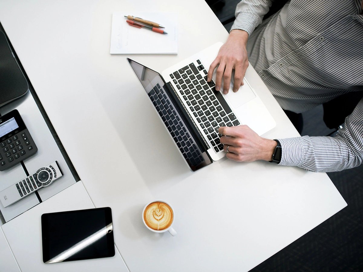 Les mains d'une personne tapent sur un ordinateur portable à un bureau avec une tasse de café et une tablette à côté.