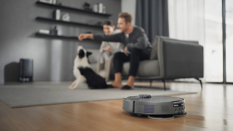Ein Saugroboter auf dem Holzboden; ein Paar interagiert mit ihrem Hund in einem modernen Wohnzimmer. Ein Saugroboter auf dem Holzboden; ein Paar interagiert mit ihrem Hund in einem modernen Wohnzimmer.