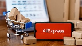 AliExpress-Logo auf einem Smartphone mit einem Einkaufswagen und Kartons im Hintergrund.