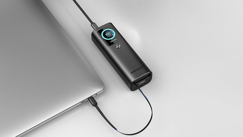 Ein schwarzer asperX Powerbank, der mit einem Laptop verbunden ist, zeigt einen Ladezustand von 100% an. Ein schwarzer asperX Powerbank, der mit einem Laptop verbunden ist, zeigt einen Ladezustand von 100% an.