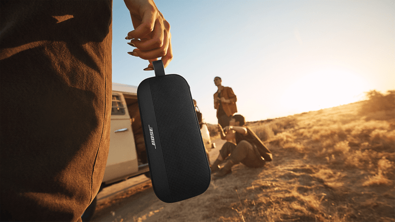 Eine Person hält einen Bose SoundLink Flex Lautsprecher, während zwei Personen im Hintergrund bei Sonnenuntergang sind.