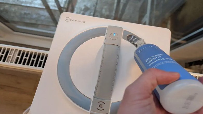 Eine Hand hält eine Flasche ECOVACS WINBOT Reinigungsmittel neben dem ECOVACS WINBOT W2 Pro Omni. Eine Hand hält eine Flasche ECOVACS WINBOT Reinigungsmittel neben dem ECOVACS WINBOT W2 Pro Omni.