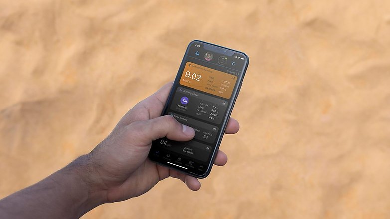 Eine Hand hält ein Smartphone, das Statistiken der Garmin Forerunner 165 Music auf einem sandigen Hintergrund anzeigt.