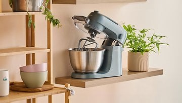 Kenwood Küchenmaschine mit einer Edelstahl-Schüssel auf einem Holzregal neben Topfpflanzen.