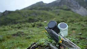 Zwei Kospet S2-Smartwatches, eine schwarze und eine weiße, auf einem Stein in einer bergigen, grasbewachsenen Landschaft.