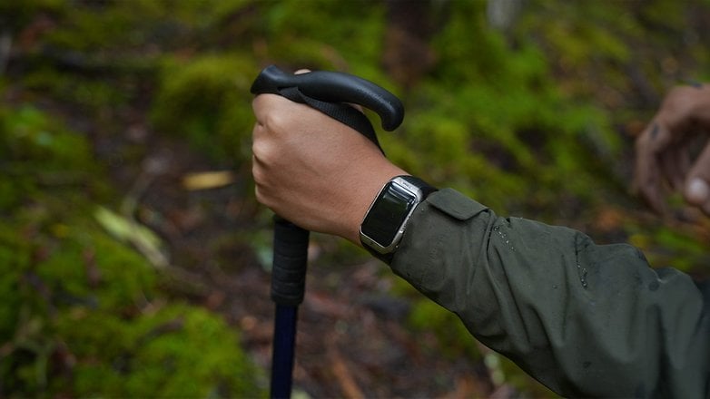 Eine Hand hält einen Wanderstock und trägt eine Kospet Tank X2 Ultra Smartwatch in einem Wald.