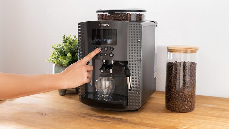 Eine KRUPS Essential EA8100 Kaffeemaschine mit einem Benutzer, der einen Knopf drückt, neben einem Glas mit Kaffeebohnen.