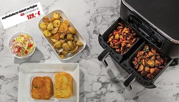 Ninja-Heißluftfritteuse mit zwei Fächern, die Gemüse und Fleisch zubereitet, daneben ein Beilagensalat und zwei Brötchen.