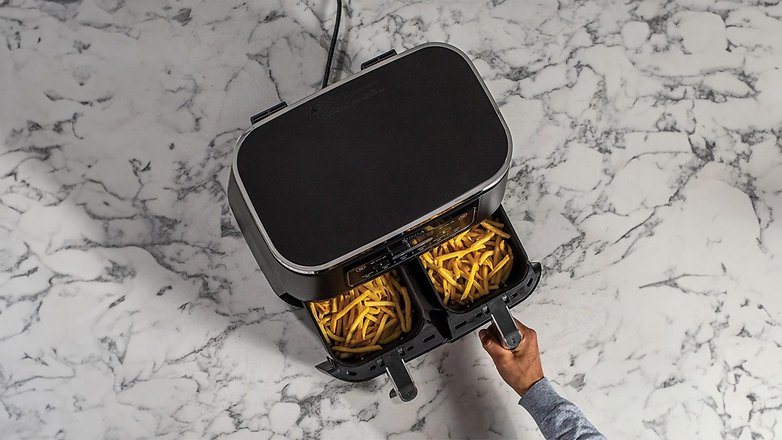 Eine Hand hält eine Schublade einer Ninja-Heißluftfritteuse, die mit goldenen Pommes auf einer Marmorplatte gefüllt ist. Eine Hand hält eine Schublade einer Ninja-Heißluftfritteuse, die mit goldenen Pommes auf einer Marmorplatte gefüllt ist.