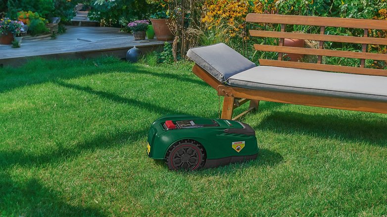 Ein grüner Mähroboter auf Gras neben einer Holzliege.