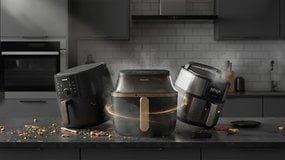 Drei Heißluftfritteusen auf einer Küchentheke, darunter ein Philips- und ein Ninja-Modell, mit verstreuten Kochzutaten.