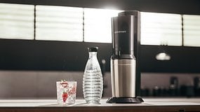 Eine SodaStream Crystal 3.0-Maschine neben einer Sprudelwasserflasche und einem Glas mit Eis und Erdbeeren.