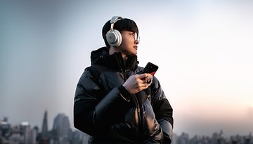 Person trägt SteelSeries Arctis Nova Elite Gaming-Headset und hält ein Smartphone vor einer Stadtsilhouette.