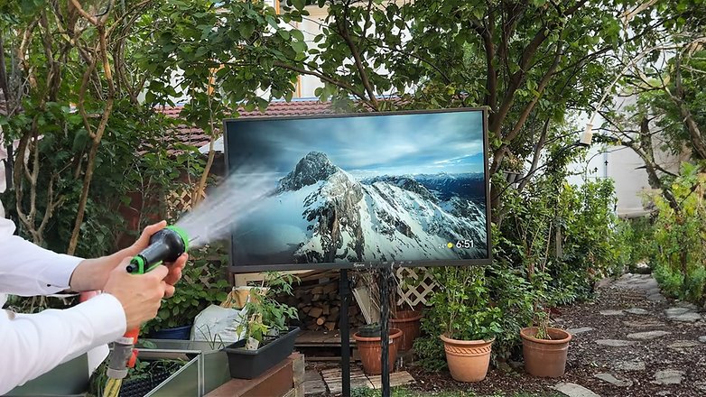 Eine Person gießt Pflanzen mit einem Schlauch, während ein Sylvox Deck Pro 2 Fernseher eine Berglandschaft zeigt.