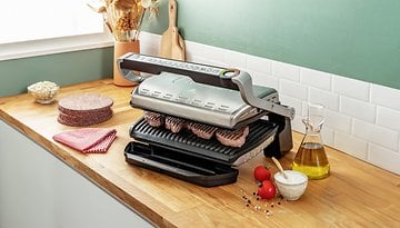 Ein Tefal Optigrill XL Plus grillt Fleisch auf einer Holzarbeitsplatte mit Gewürzen und Zutaten in der Nähe.