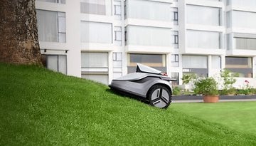 Ein Mähroboter auf einer geneigten Grasfläche neben einem Baum und modernen Gebäuden im Hintergrund.