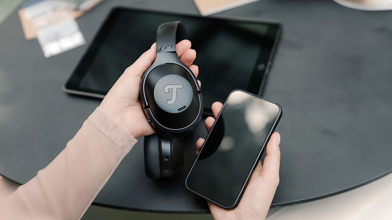 Eine Person hält schwarze Teufel Over-Ear-Kopfhörer und ein Smartphone auf einem Tisch. Eine Person hält schwarze Teufel Over-Ear-Kopfhörer und ein Smartphone auf einem Tisch.