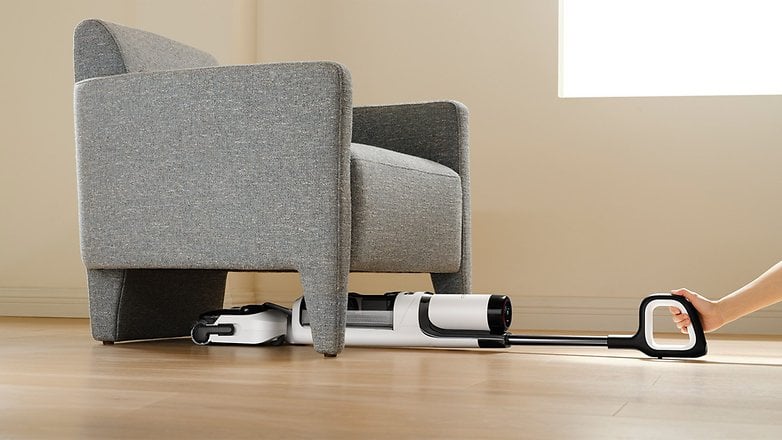 Ein Tineco Floor One S6 Staubsauger wird unter einem grauen Sessel herausgezogen. Ein Tineco Floor One S6 Staubsauger wird unter einem grauen Sessel herausgezogen.