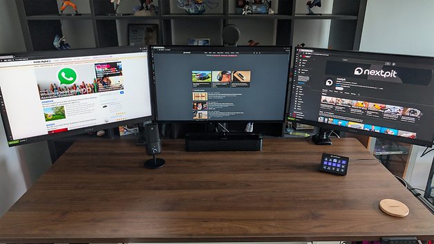 Ein moderner Arbeitsplatz mit drei Monitoren auf einem Holztisch, die verschiedene Websites und Apps anzeigen.