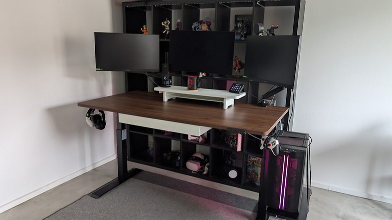 Ein modernes Schreibtisch-Setup mit einem großen Holztisch, drei Monitoren, Gaming-Zubehör und einem Regal zur Aufbewahrung. Ein modernes Schreibtisch-Setup mit einem großen Holztisch, drei Monitoren, Gaming-Zubehör und einem Regal zur Aufbewahrung.