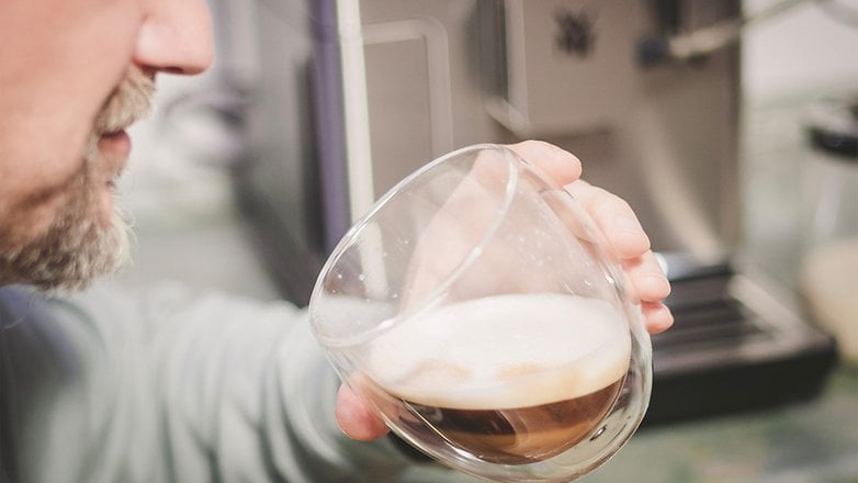 Eine Person hält ein Glas mit Kaffee und Schaum vor einer Kaffeemaschine.