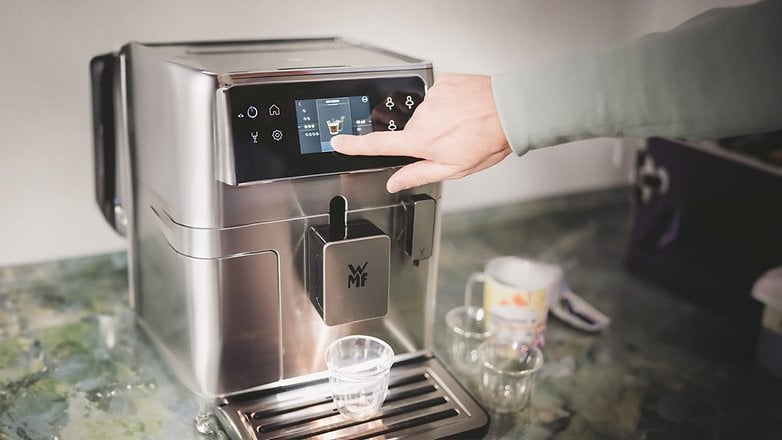 Eine WMF-Kaffeemaschine mit Touchscreen, eine Hand, die Einstellungen anpasst, und einem Glas darunter.