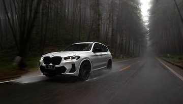 A white BMW X3 driving on a wet road through a foggy forest.