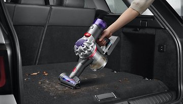 Eine Person benutzt einen Dyson V8 kabellosen Staubsauger, um den Kofferraum eines Autos mit Schmutz und Ablagerungen zu reinigen.