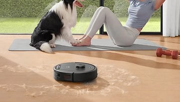 A robotic vacuum cleaner is cleaning up pet hair on a wooden floor while a woman exercises with a dog nearby.