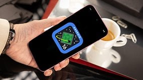 Une main tenant un smartphone affichant le logo Android 16 sur une table avec une tasse de café.