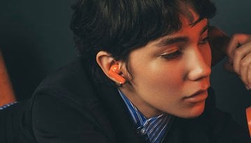A person with short hair wearing orange earbuds, looking thoughtfully to the side.