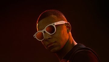 A person wearing white sunglasses against a dark background.